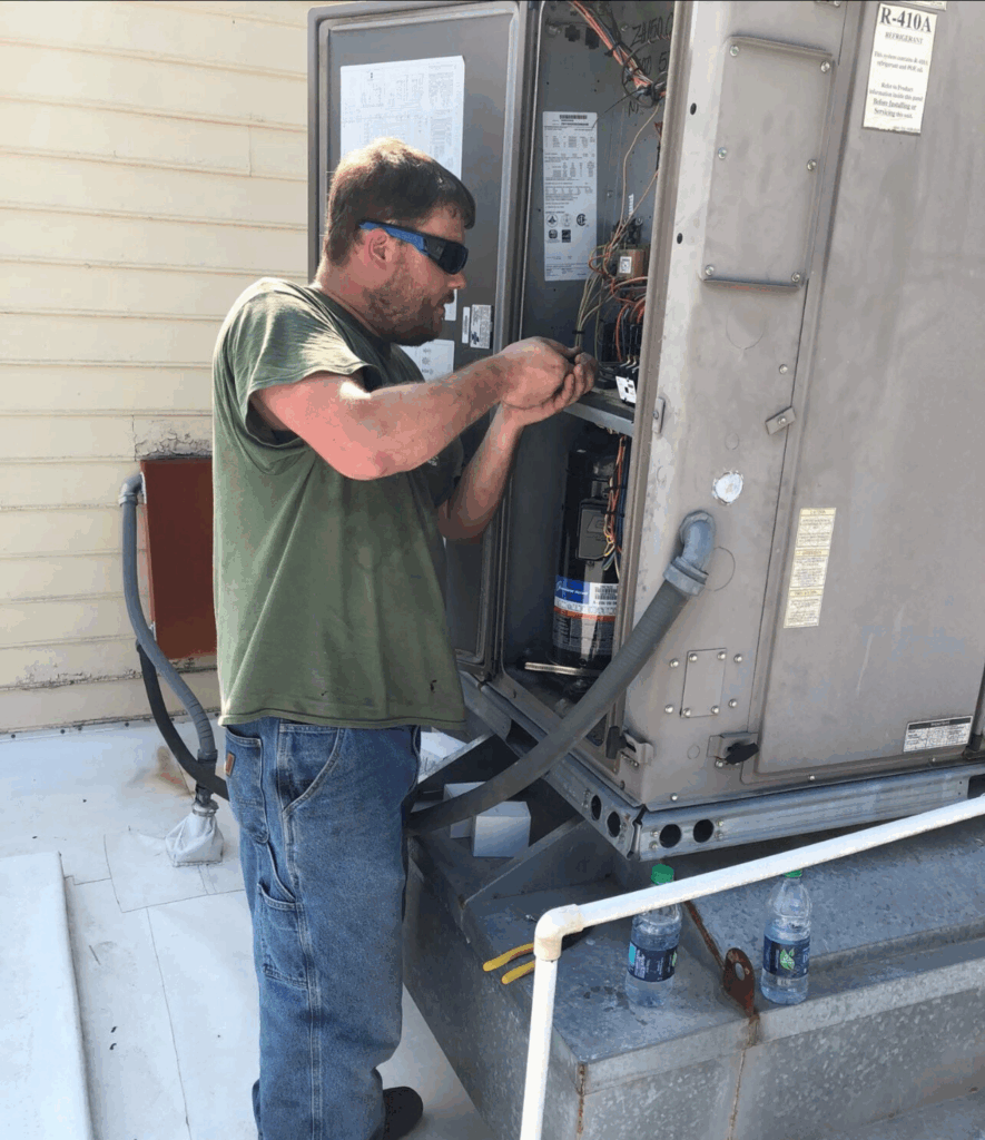 Man in sunglasses and green shirt repairs wiring inside an outdoor HVAC unit on a rooftop.