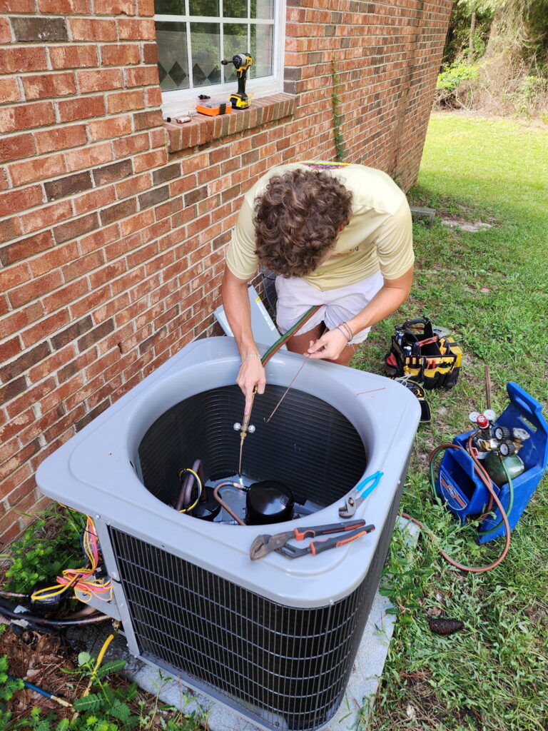 A man repairs an outdoor air conditioning unit next to a brick house, using tools and gauges.